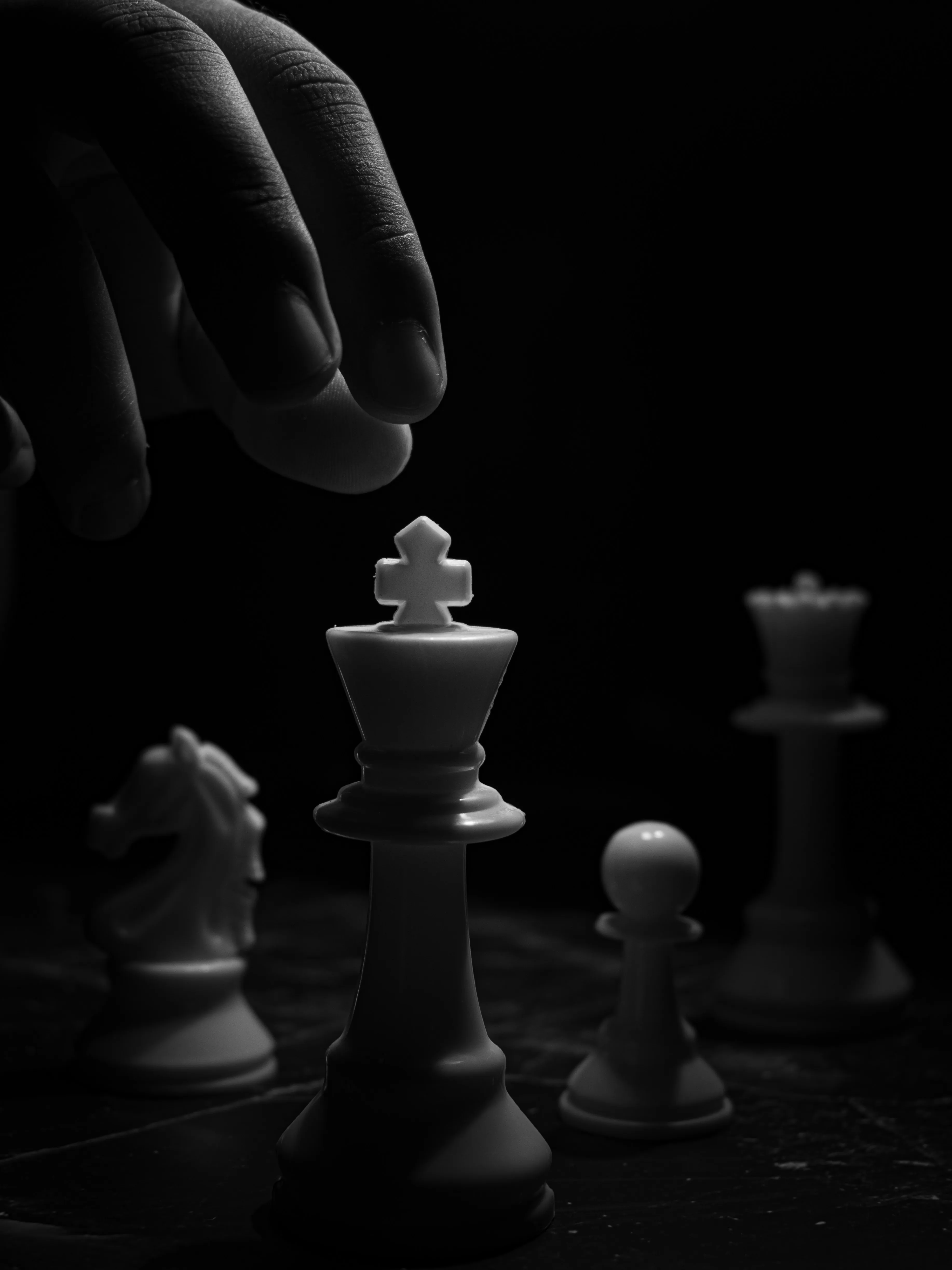 Hand about to move a white king chess piece with a knight, pawn, and queen in black background.