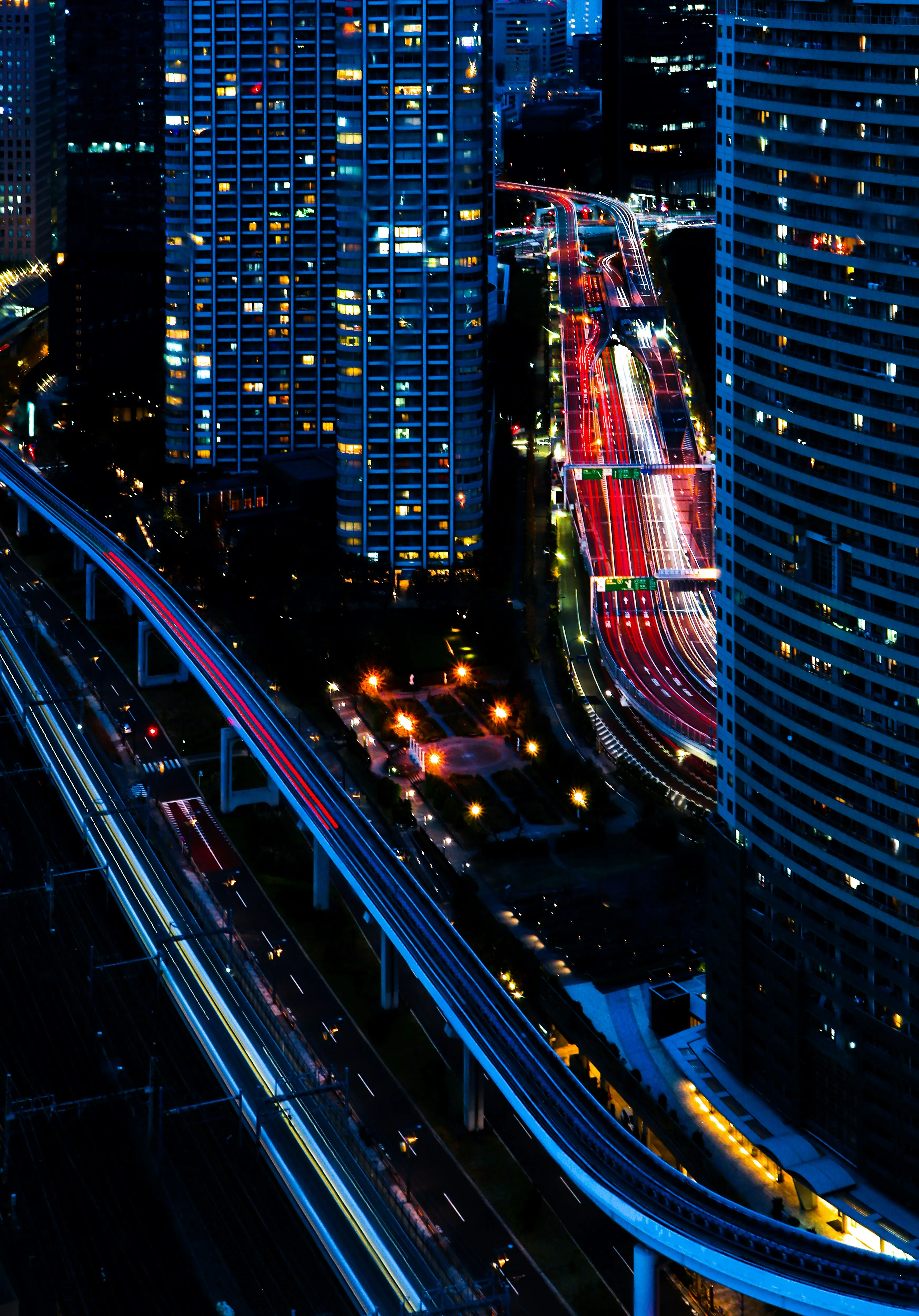 Night aerial view of city highway and rail tracks with light trails from passing vehicles and illuminated buildings.