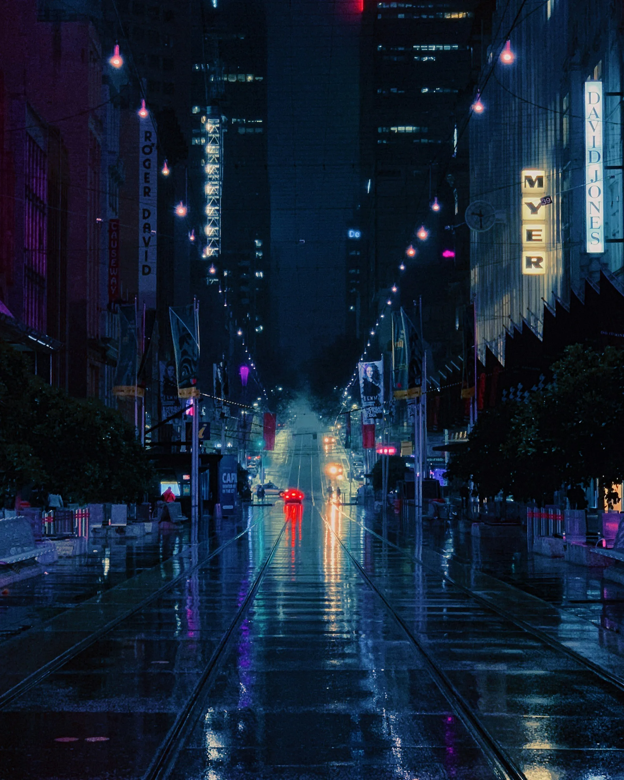 Wet city street at night with tram tracks, glowing lights, and reflections on the pavement.