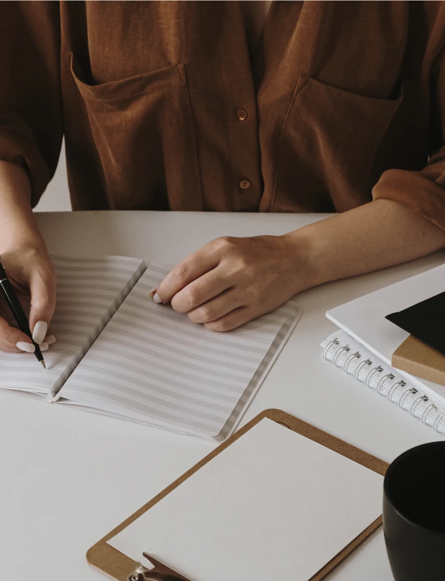 Person in brown shirt writing in a striped notebook at a white desk with papers and a black cup.