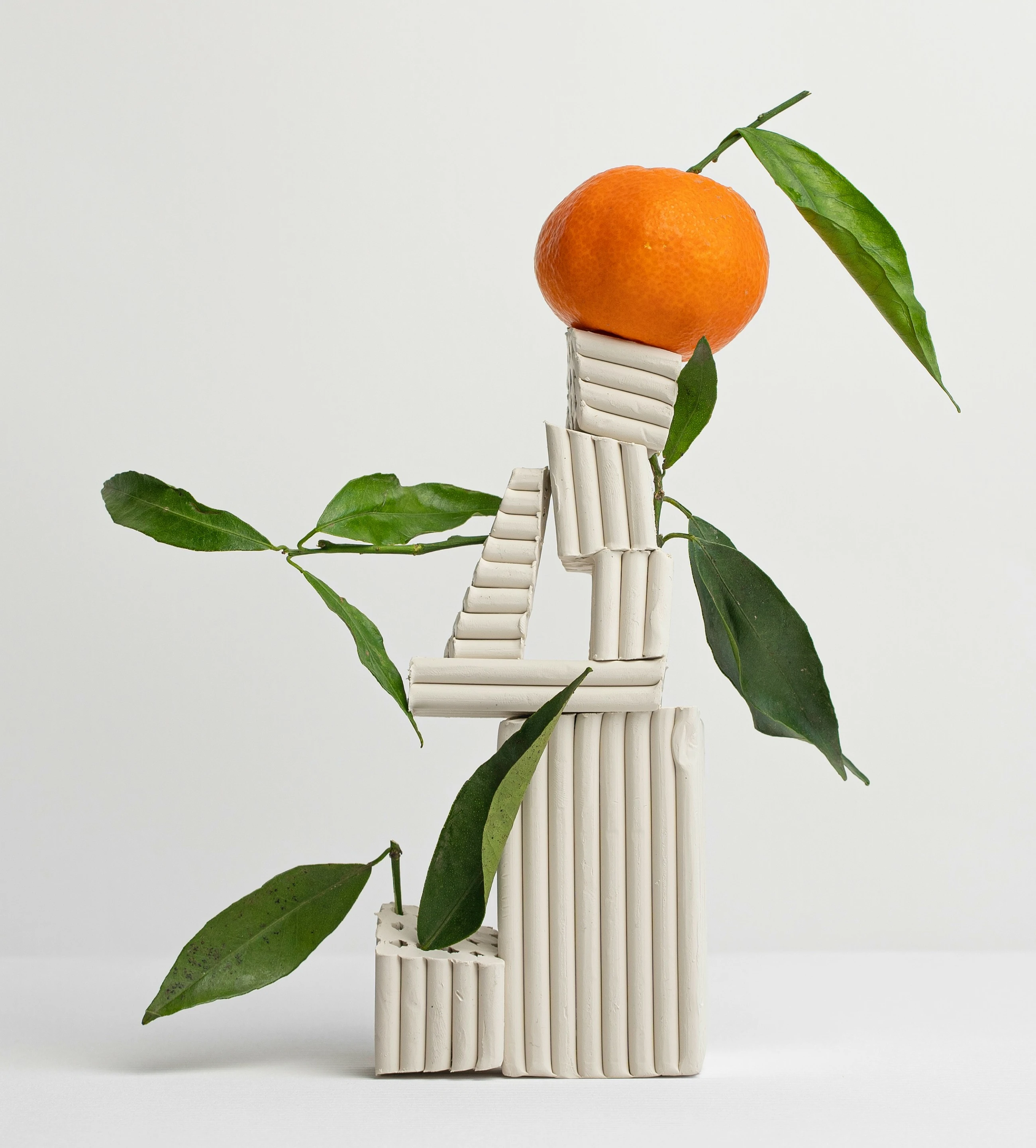 Abstract sculpture made of clay and green leaves topped with an orange fruit