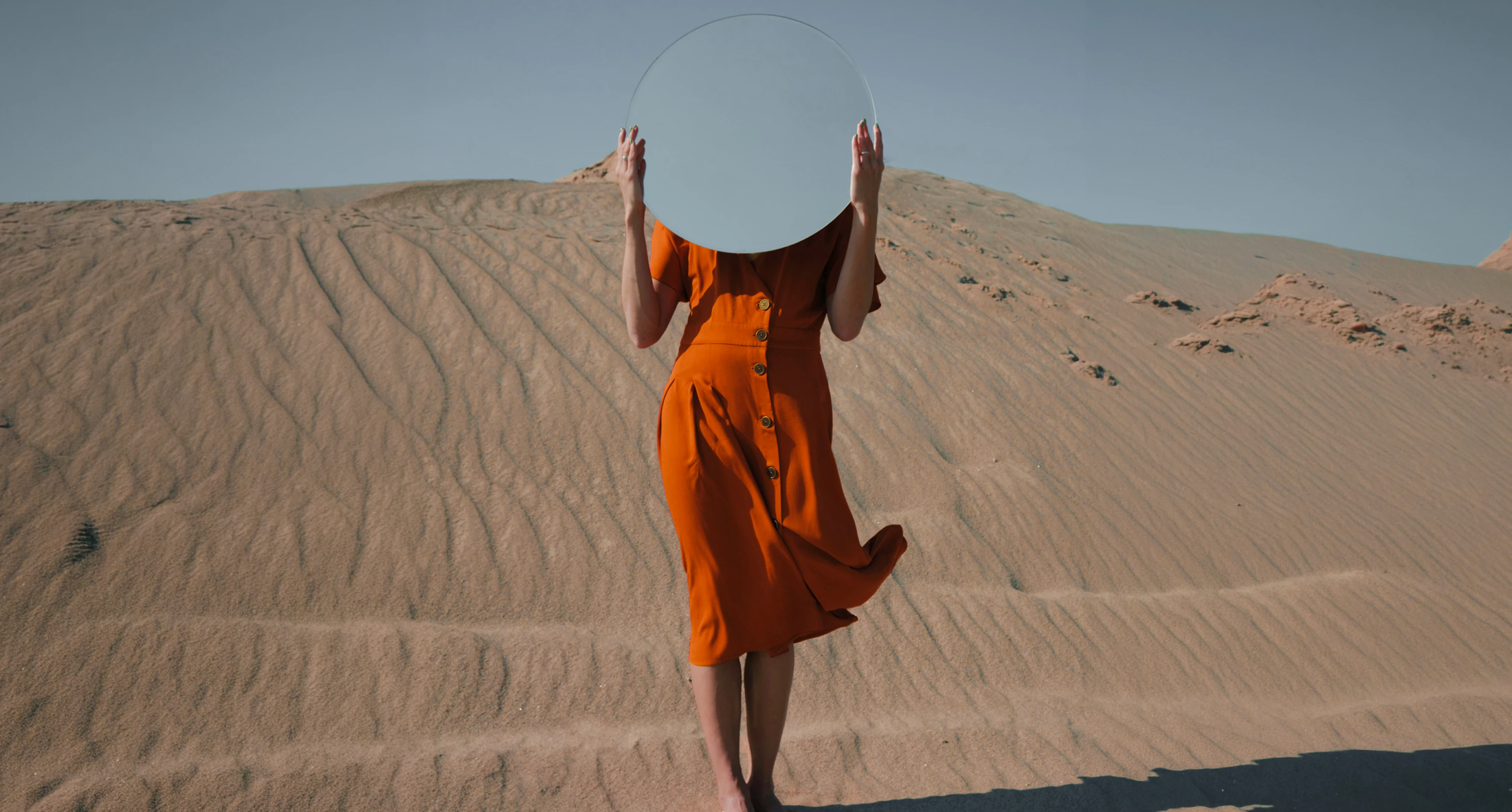 Person holding a round mirror in front of their face standing on sandy ground.
