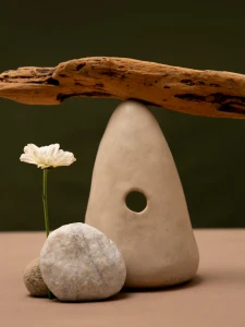 White flower next to smooth stones and a ceramic shape with a hole, holding a piece of driftwood.