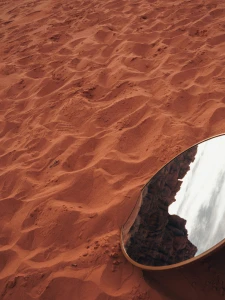Mirror on orange-red sand reflecting a rocky formation and cloudy sky.