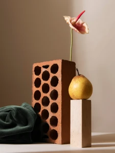 Still life with a brick, a single flower, a pear, and dark green fabric on a light surface.