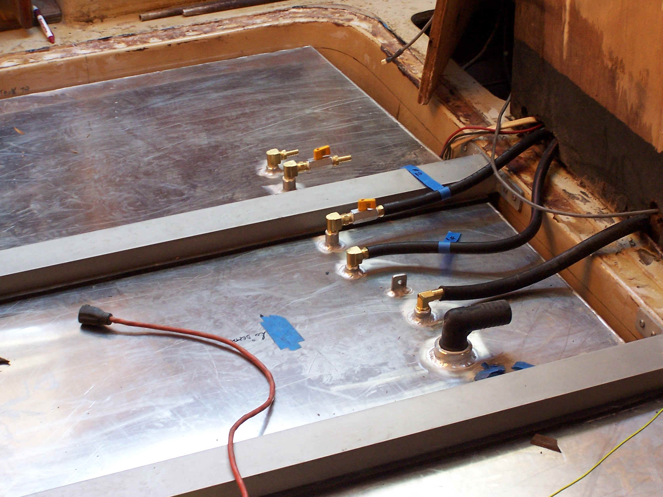 Two aluminum fuel tanks fitted into a boat's hull during a refit, with brass fittings, black fuel hoses and a red bonding wire connected on top.