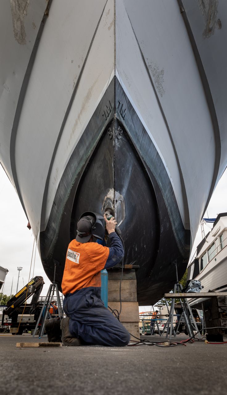 A technician in an orange jacket and ear protection kneeling beneath a hauled-out boat to sand the black antifouling paint on its keel in a boatyard.