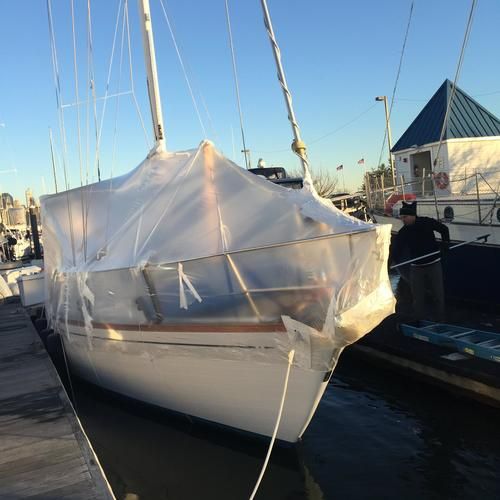 A sailboat shrink-wrapped in white plastic for winter storage while moored at a dock, with a worker standing alongside.
