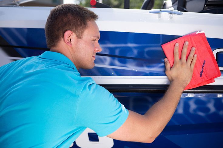 A technician in a teal polo shirt polishing the blue and white hull of a boat with a red microfiber cloth.