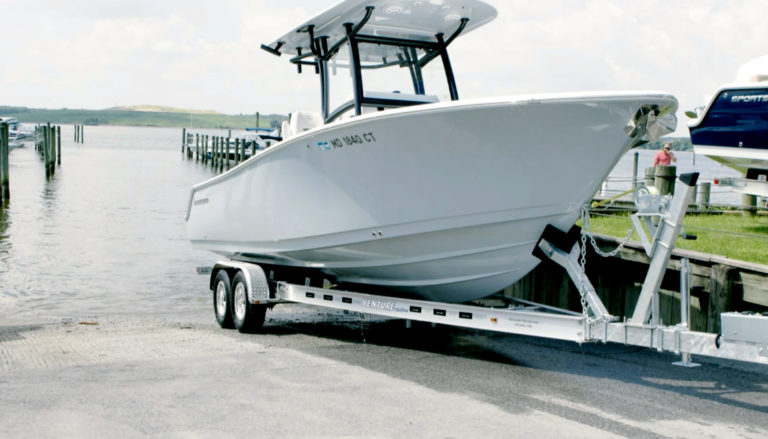 White center-console boat with a T-top sitting on a twin-axle aluminum trailer beside a wooden dock