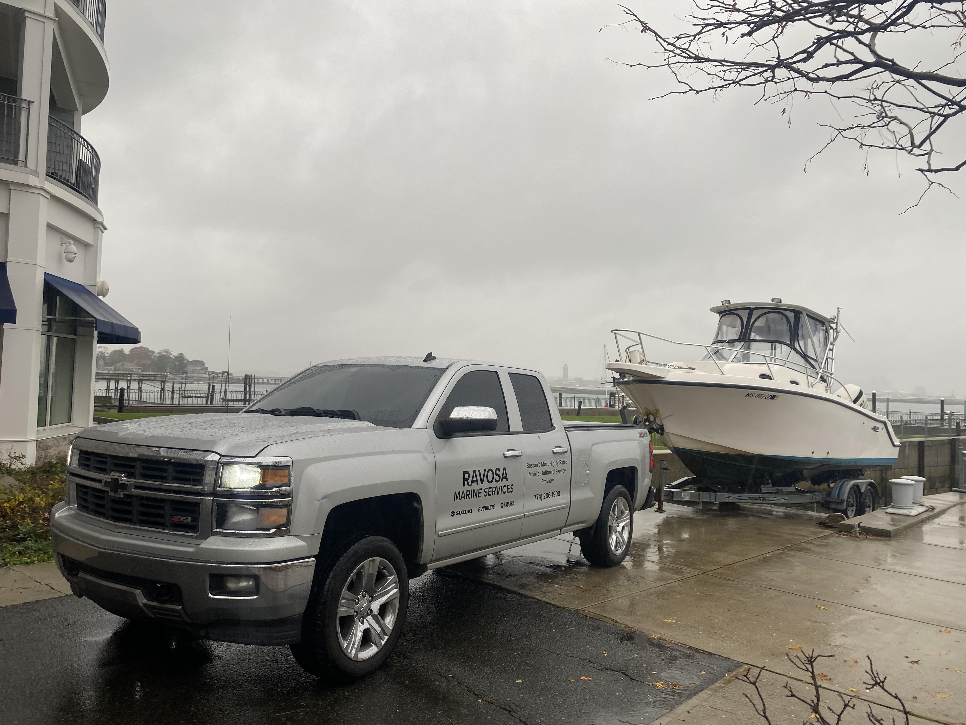 A silver pickup truck lettered Ravosa Marine Service towing a white sportfishing boat on a trailer along a waterfront driveway on an overcast day.