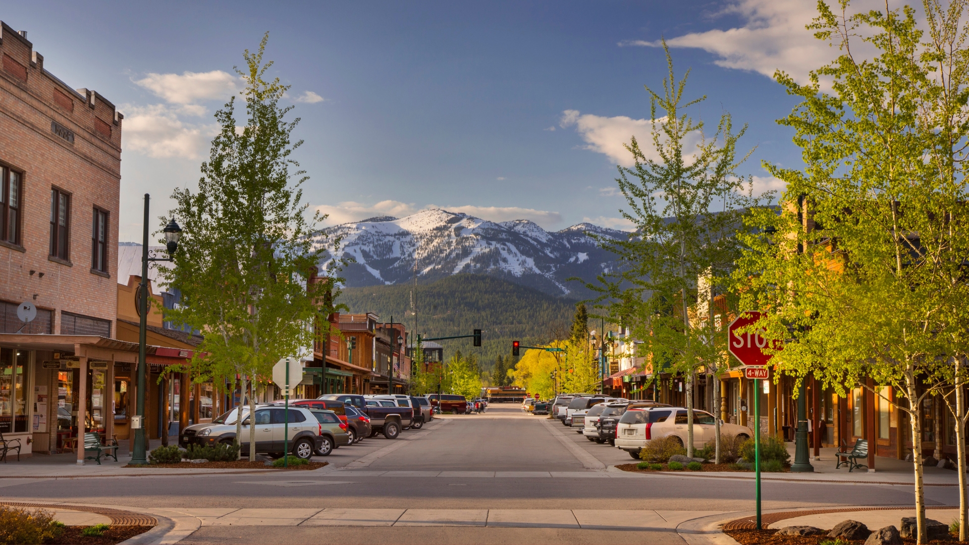 Whitefish main street with parked cars, green trees, and snow-capped mountains under a partly cloudy sky.