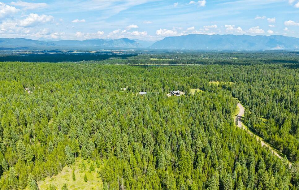 Aerial view of Whitefish Hills with dense green forest with scattered houses and mountains in the background under blue sky.