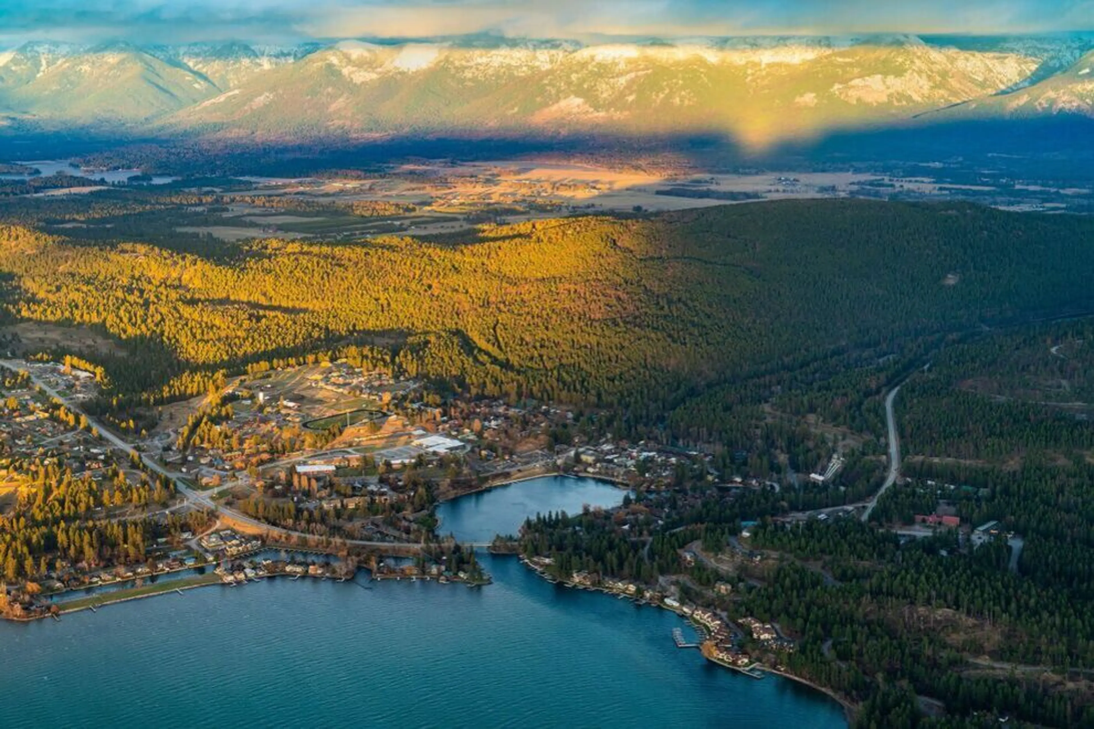 Aerial view of Bigfork , Montana, bordered by dense forest and mountains in sunlight and shade.