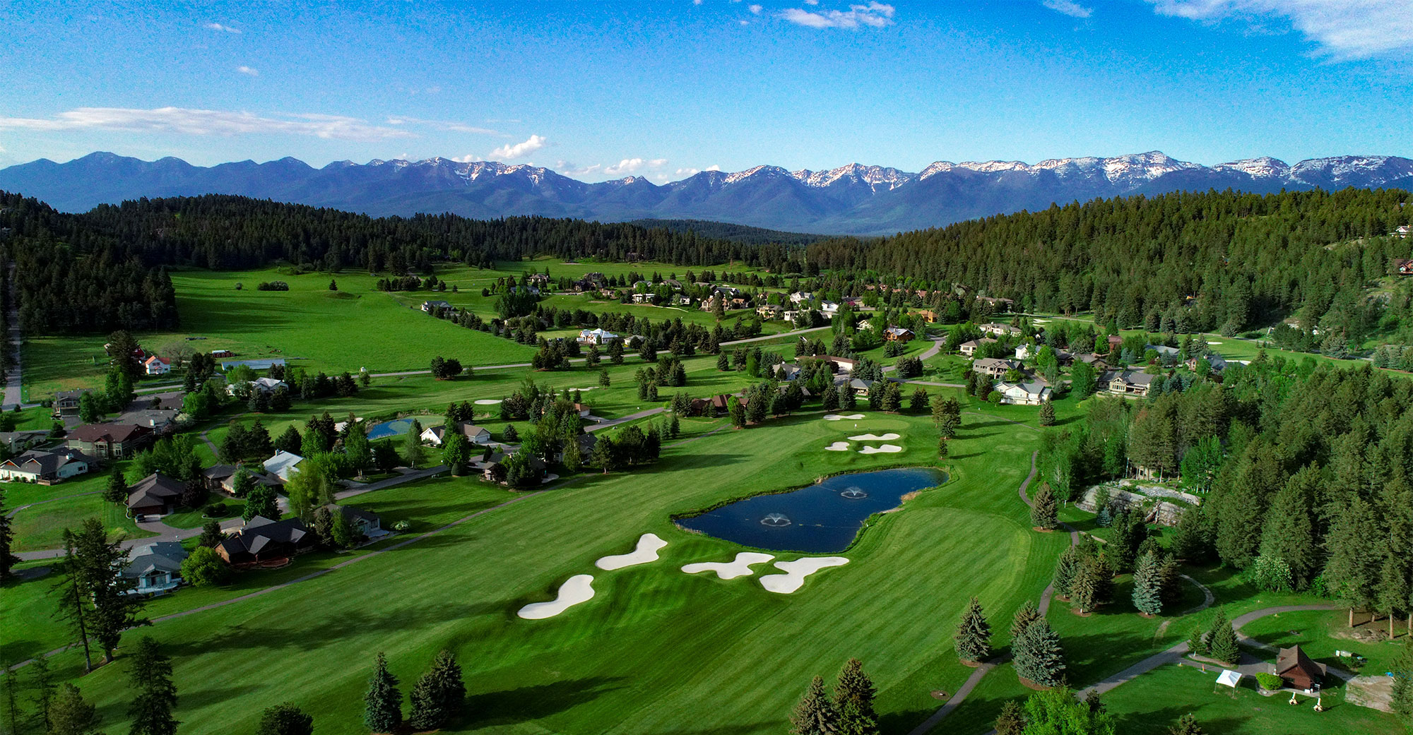 Aerial view of Eagle Bend golf course with sand traps, ponds, homes, forest, and snow-capped mountains in the background. Located in Bigfork, Montana.