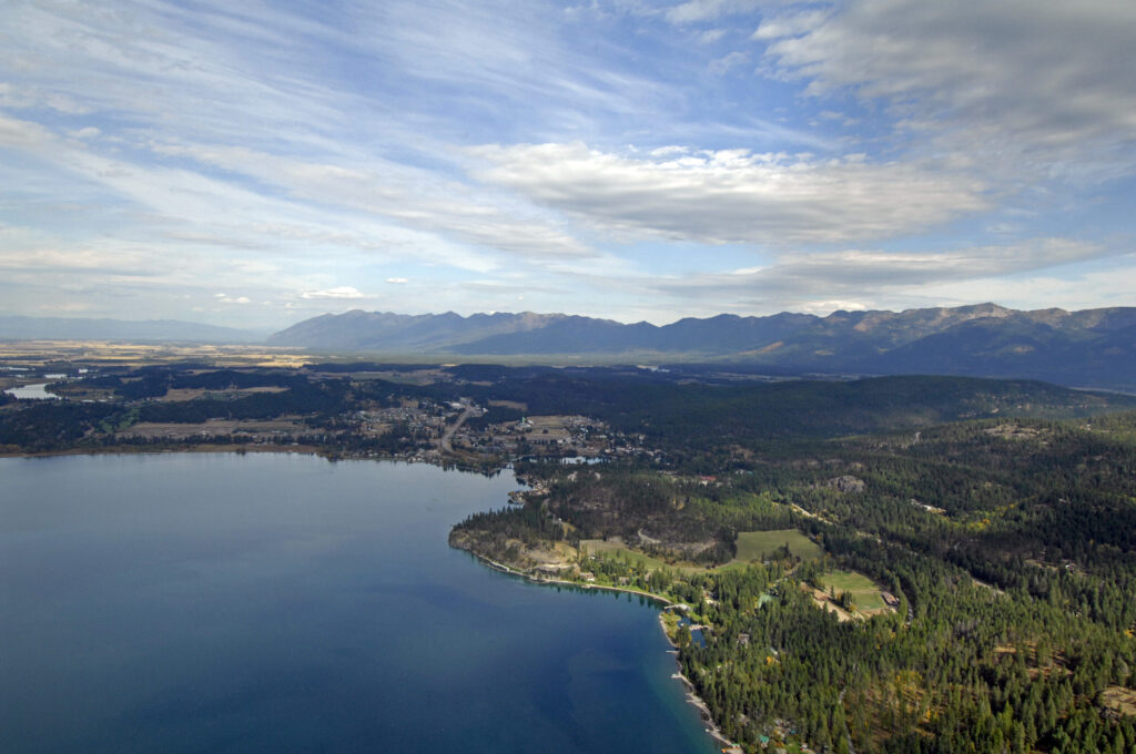Aerial view of Flathead Lake, bordered by trees with distant mountains beneath a partly cloudy sky.