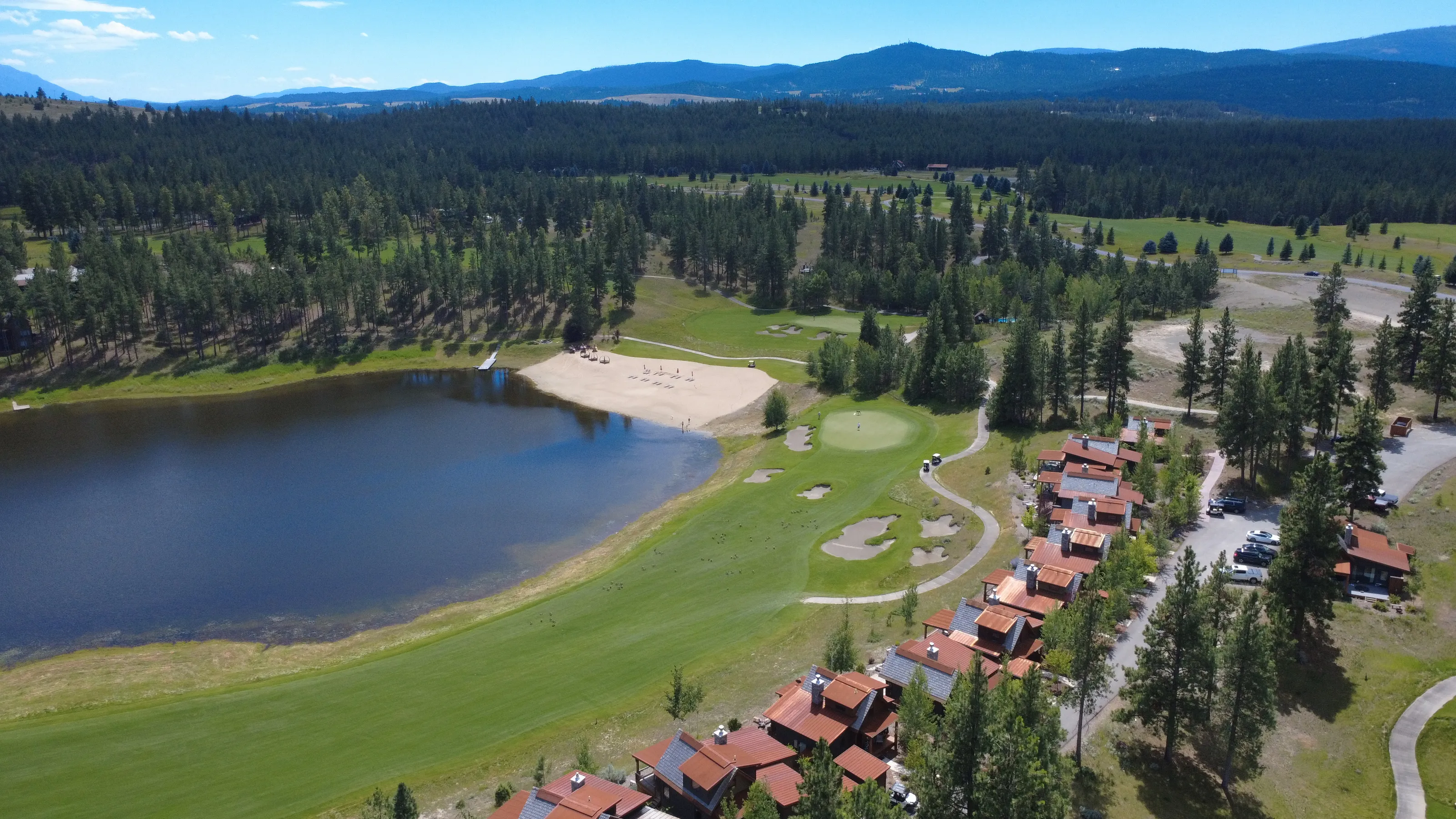 Aerial view of a lake, sandy beach, golf course, and a row of houses with reddish roofs among tall trees. Wilderness Club, Eureka, Montana.