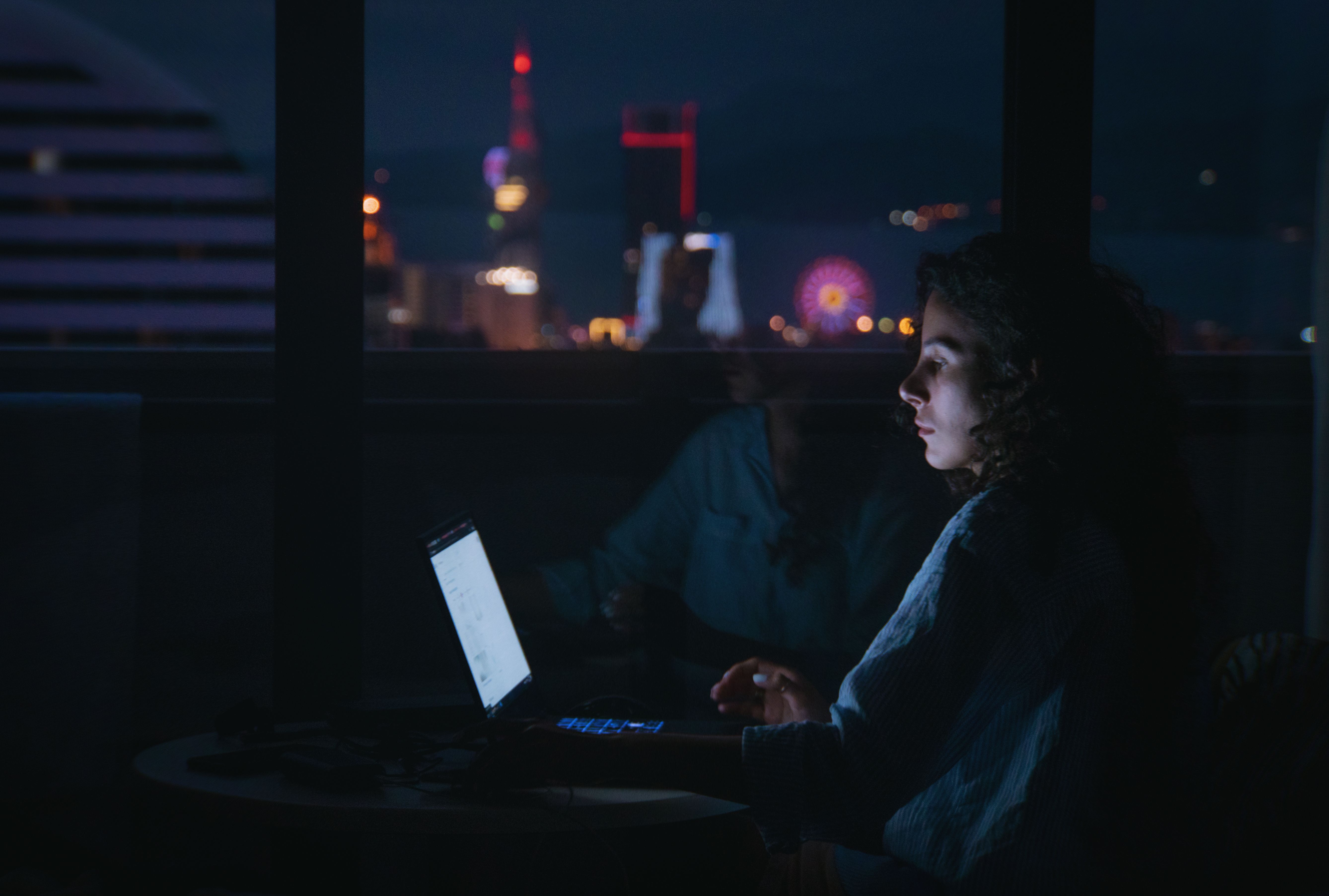 A woman focused on her laptop in a dark room with city lights visible through the window