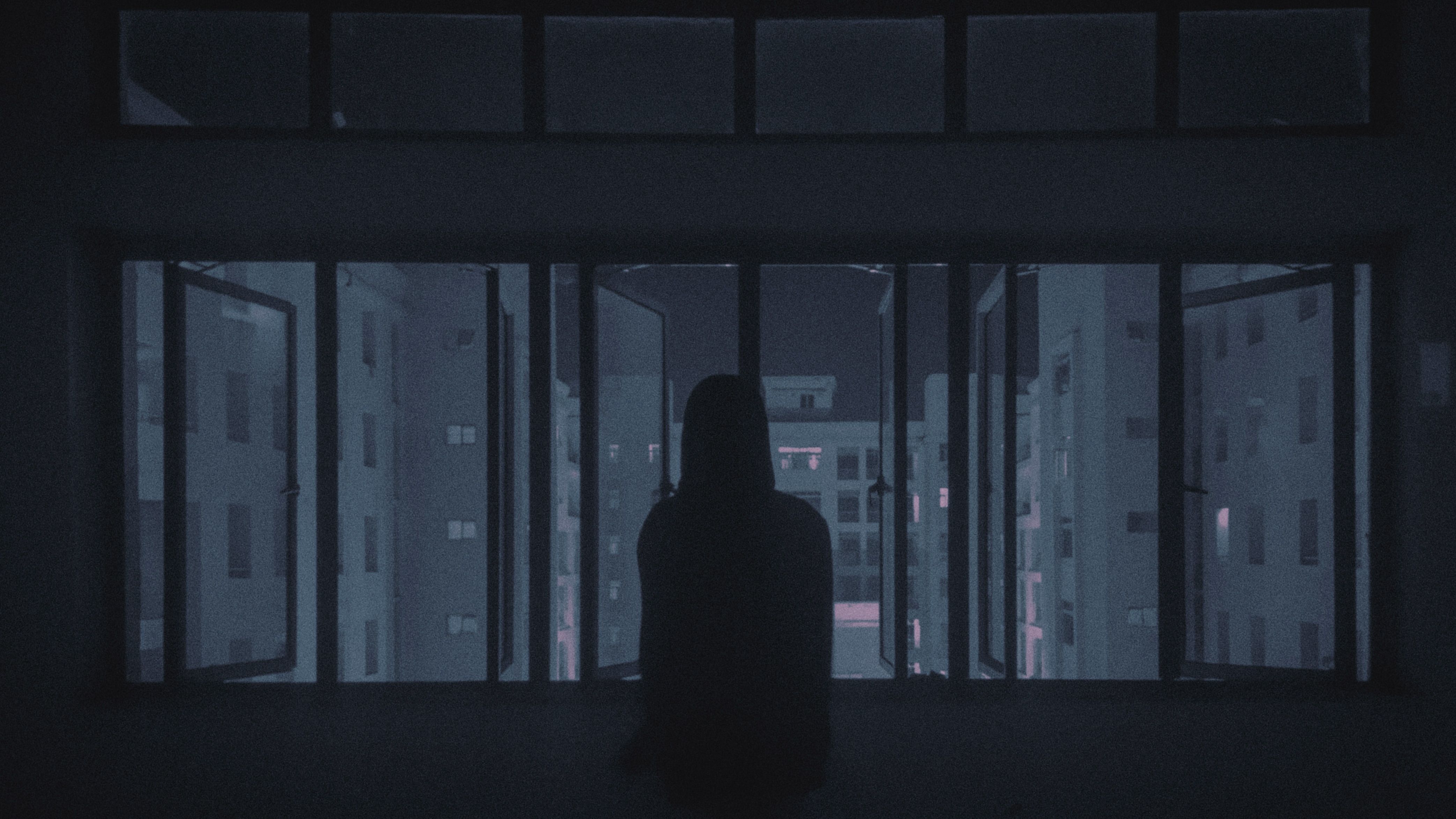 Silhouette of a person standing by a window overlooking city buildings at night.