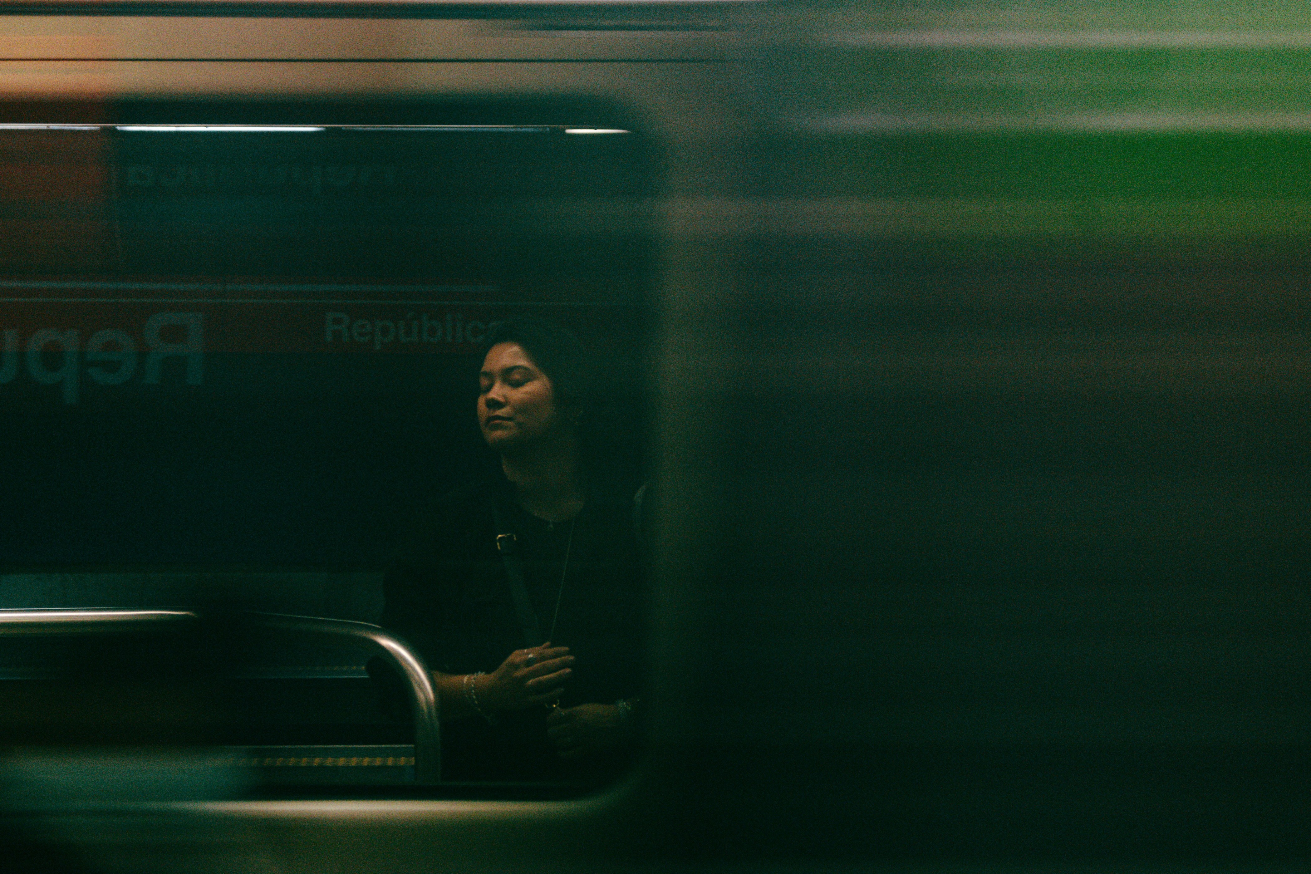 A woman stands on a subway platform with a passing train, creating a motion blur effect