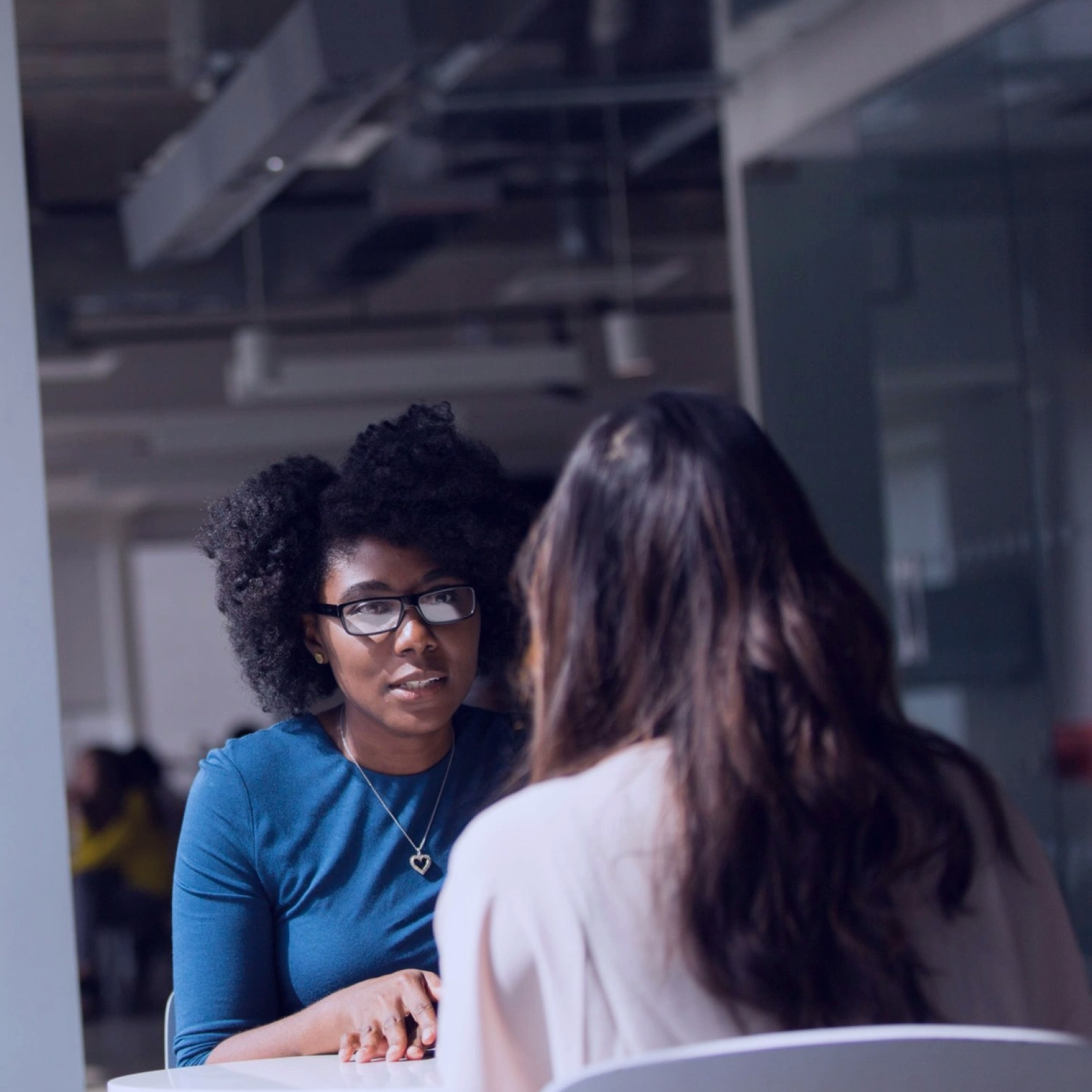 Two women having a face-to-face conversation across a table in a modern office, with one woman wearing glasses and listening attentively