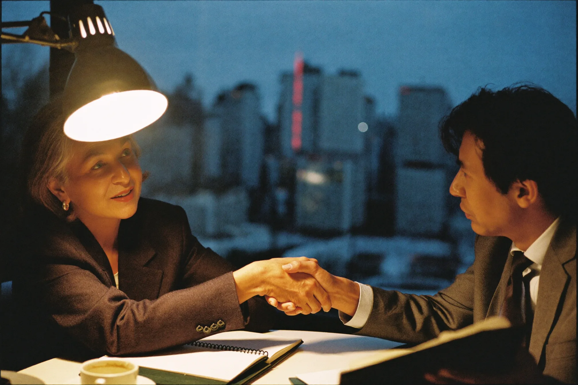 Two business people shaking hands across a small table under a warm desk lamp, dusk city skyline behind them.