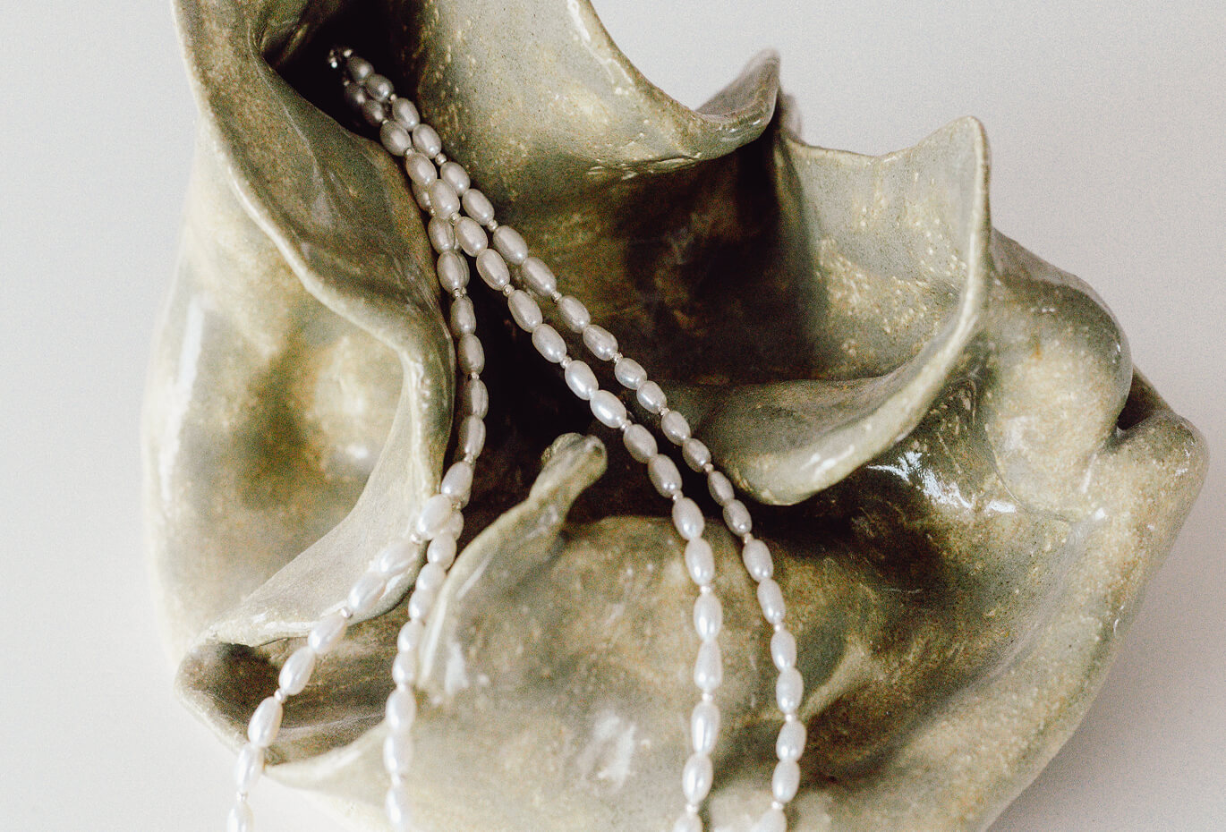 Close-up of a pearl necklace draped over a sculptural ceramic shell, combining organic texture, soft light and jewelry detail.