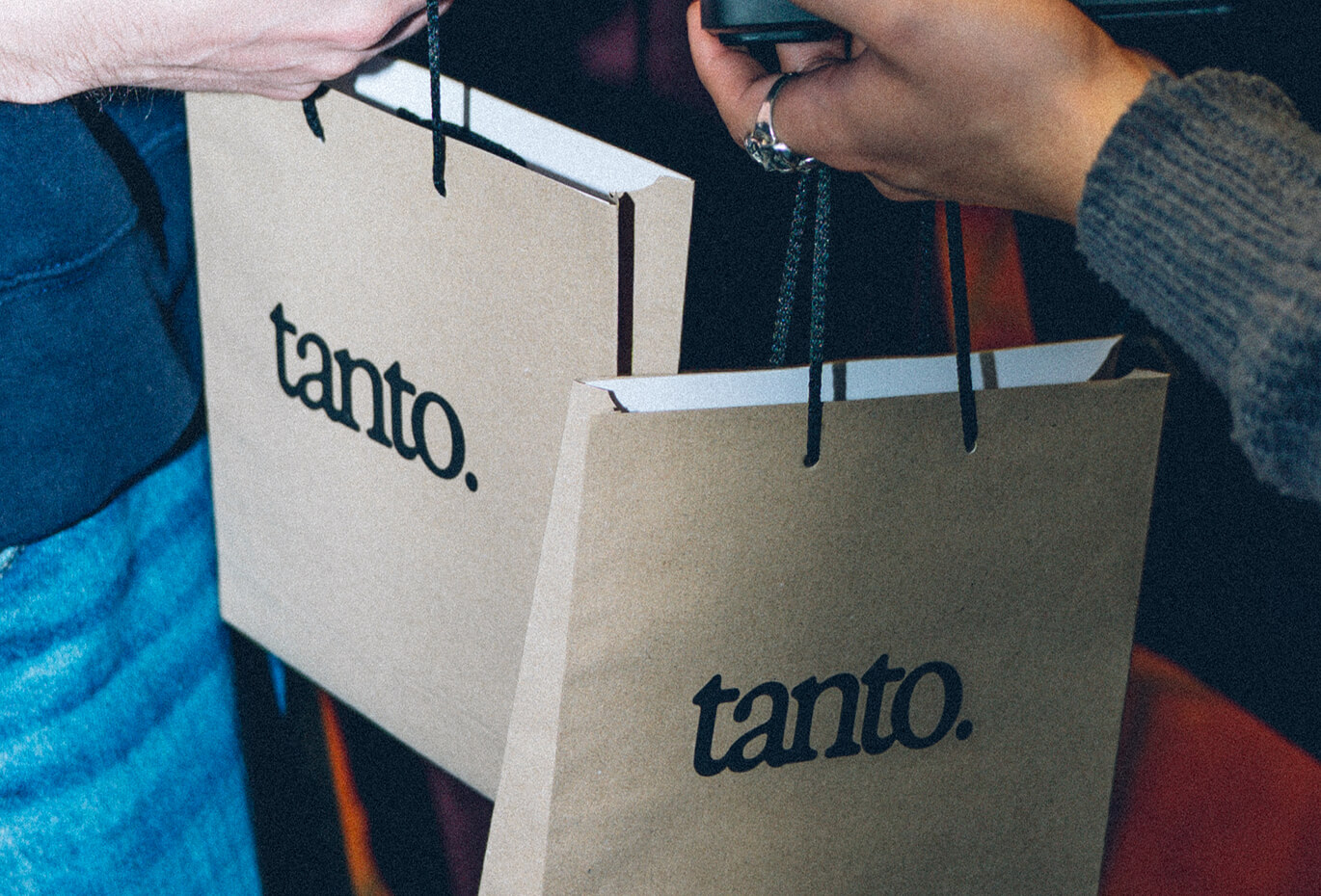Close-up of kraft paper shopping bags with the Tanto logo being carried by two people in a dark, grainy photographic style.