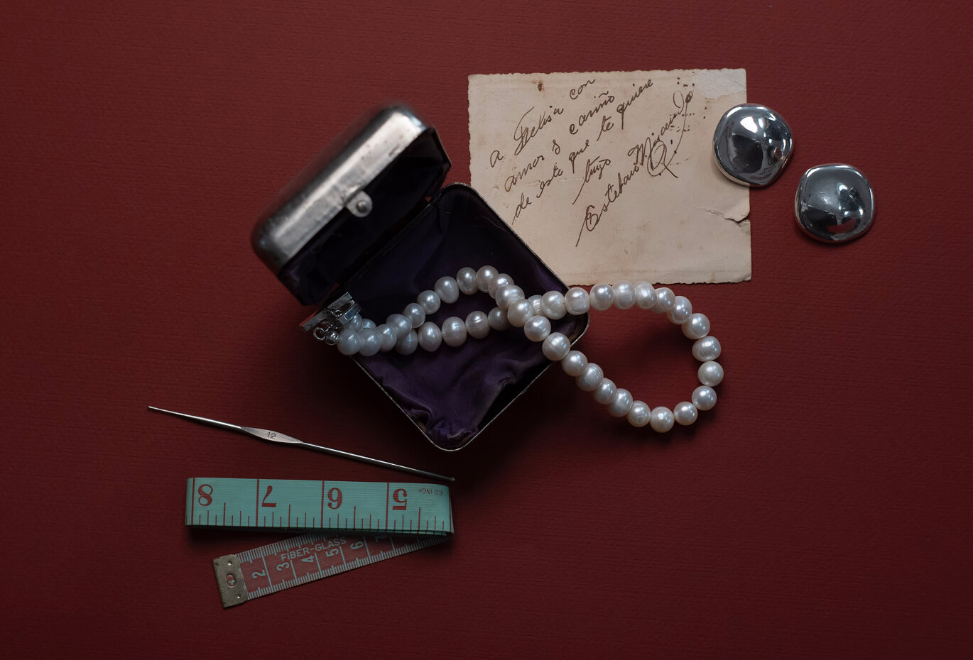 Pearl necklace arranged with vintage objects, handwritten paper, measuring tape and metallic pieces on a deep red surface.