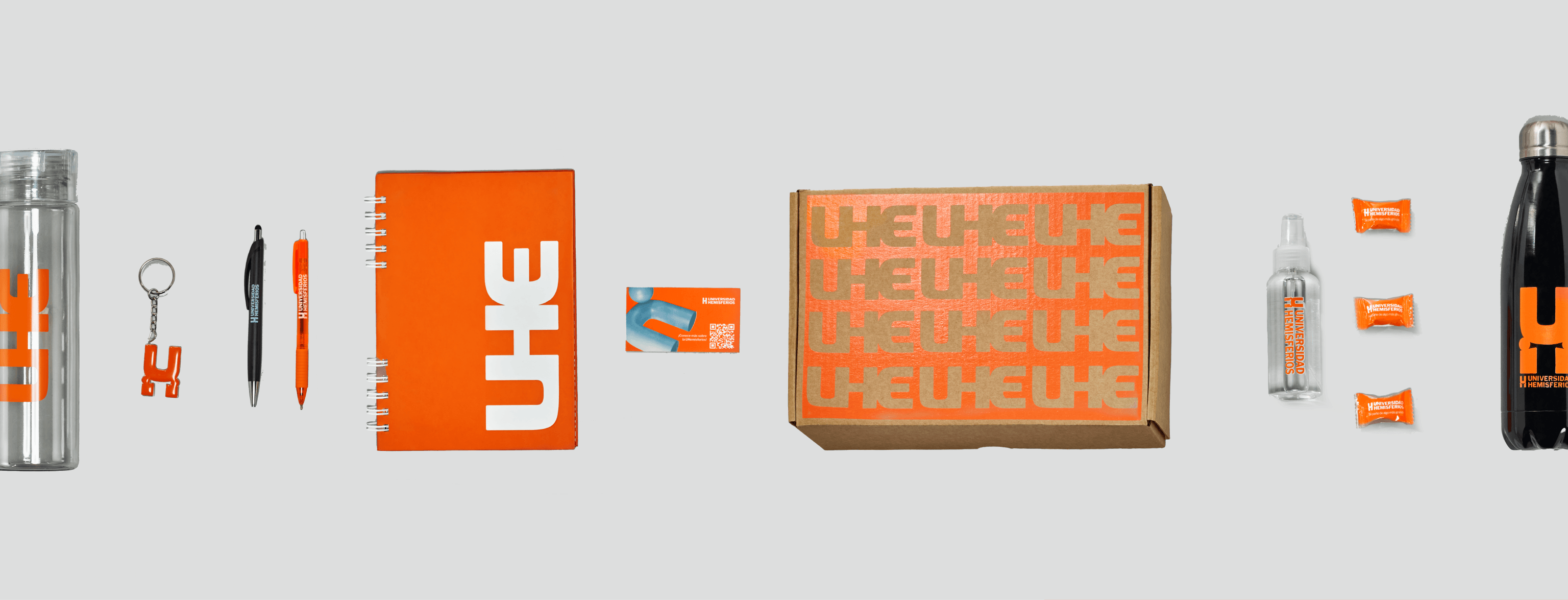 Branded merchandise kit for Universidad de los Hemisferios by Lunalunares, showing a water bottle, keychain, pens, notebook, card, box, sanitizer, candies, and reusable bottle with orange UHE applications.