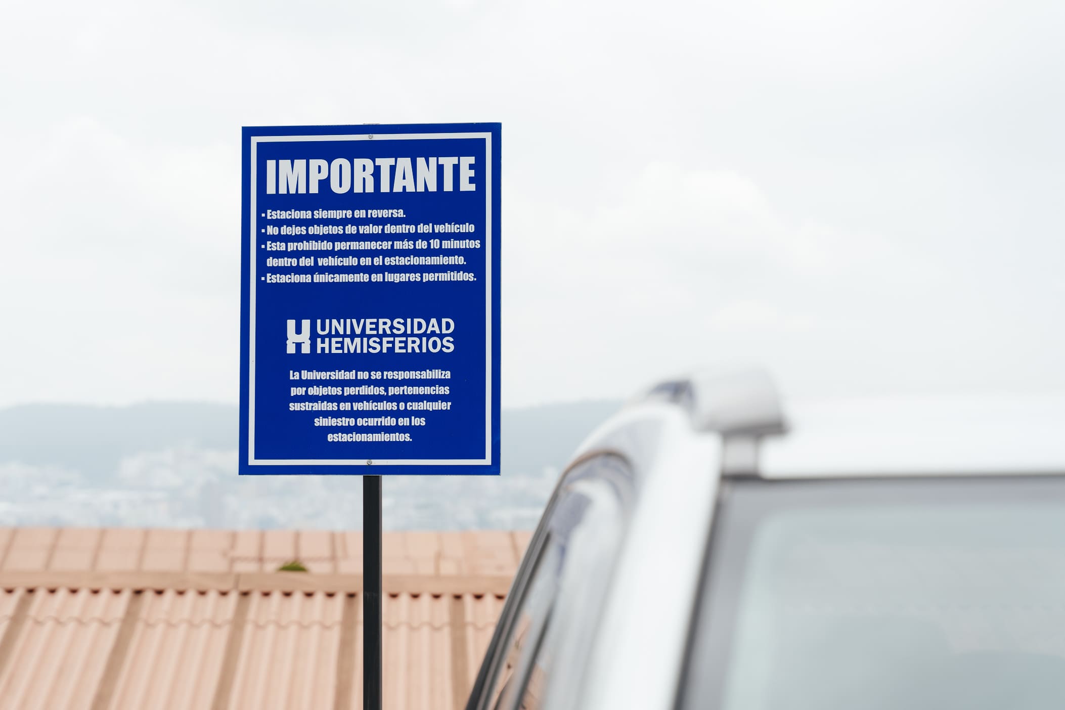 Parking signage system for Universidad de los Hemisferios by Lunalunares, featuring a blue informational sign with institutional logo, safety instructions, and outdoor campus context.