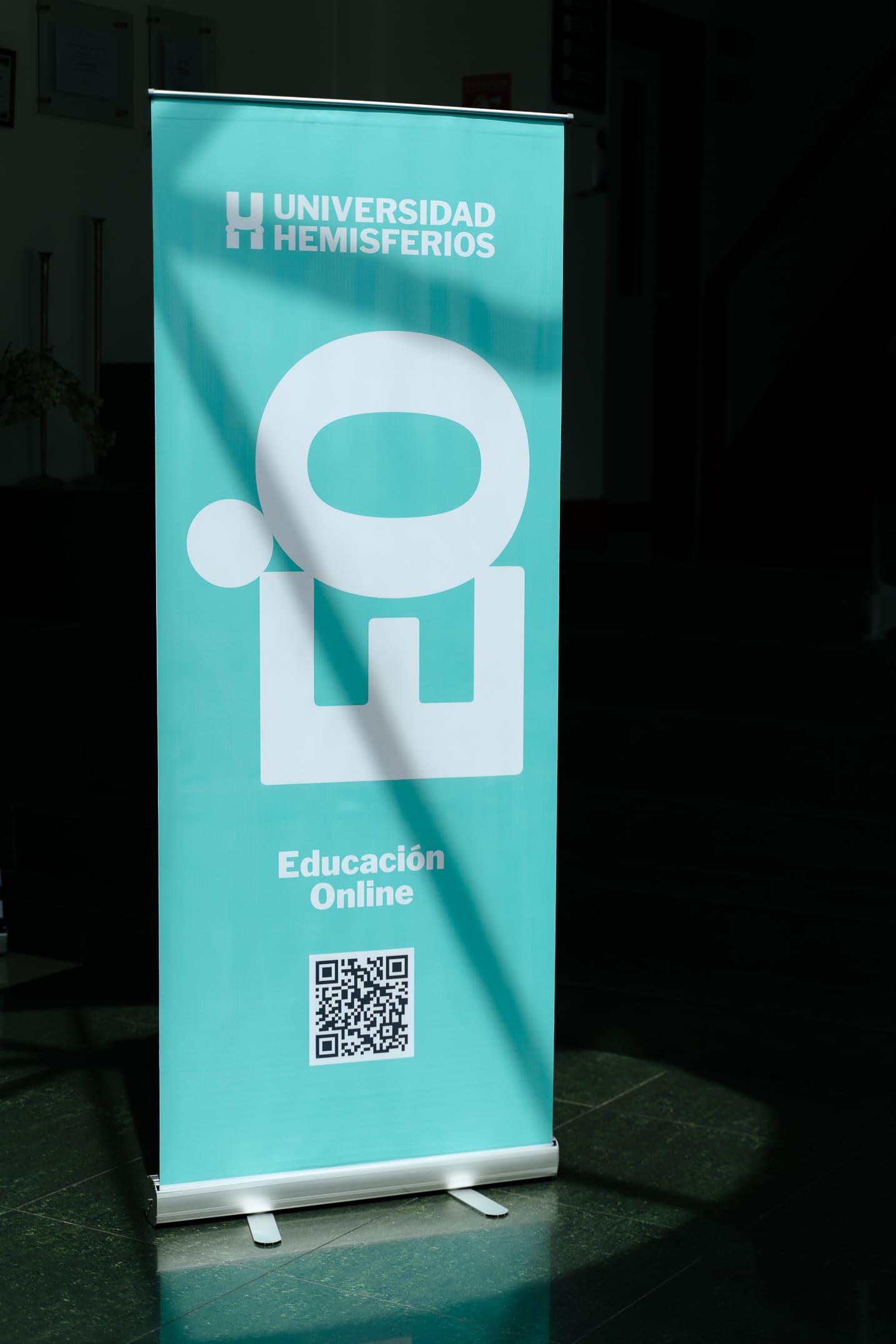 Online education banner design for Universidad de los Hemisferios by Lunalunares, featuring a turquoise roll-up display with UHE branding, white academic icon, and QR code application.