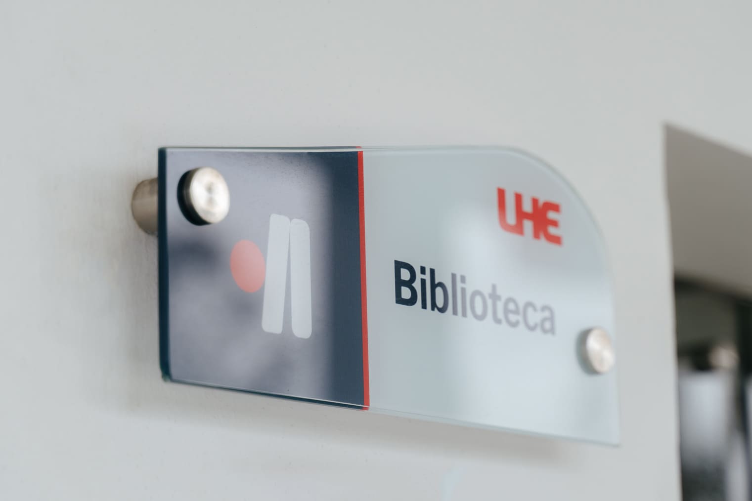 Library wayfinding signage design for U-Hemisferios by Lunalunares, showing a frosted acrylic wall sign with the Biblioteca service mark, UHE logo, and institutional color accents.