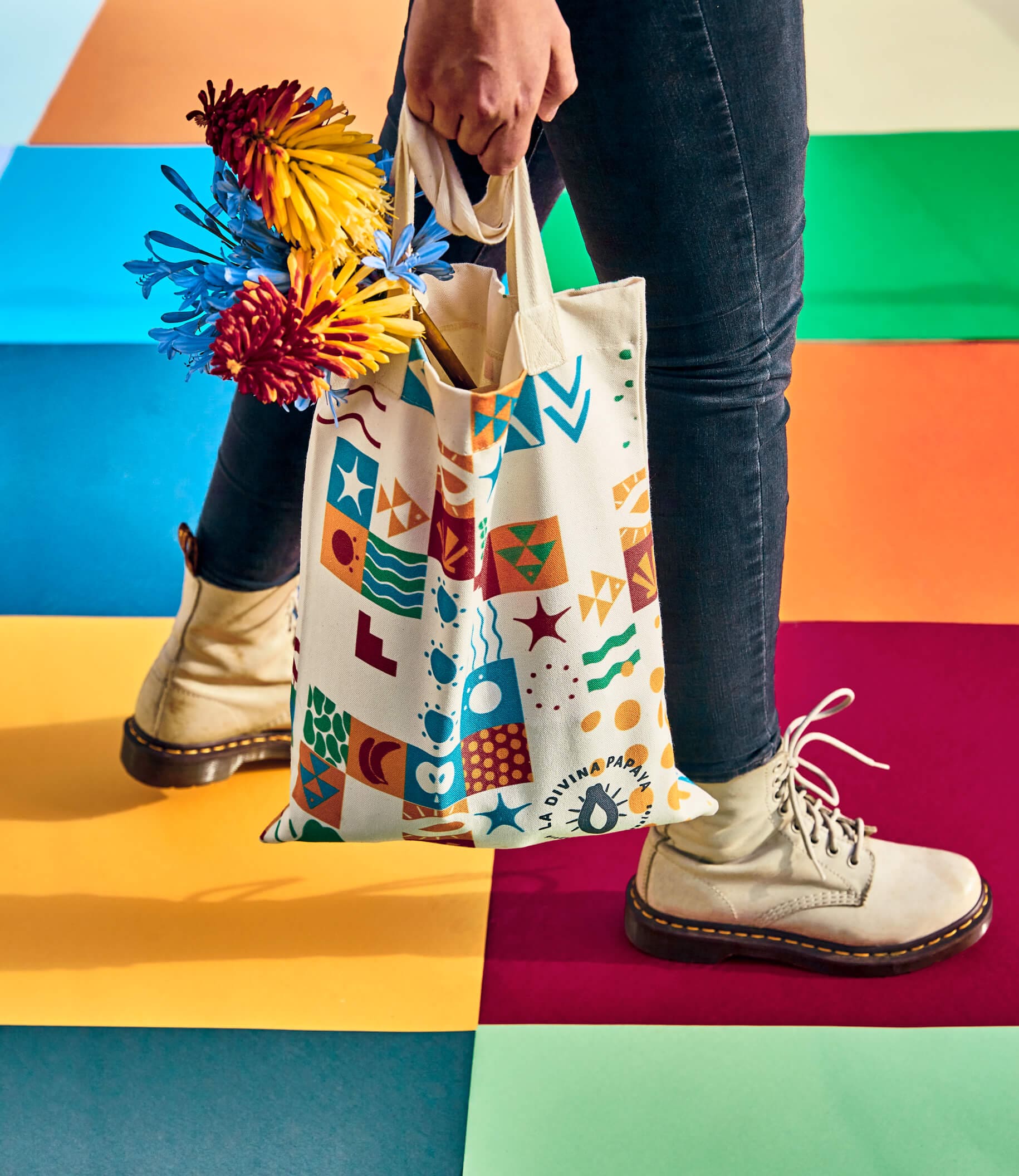 Patterned tote bag for La Divina Papaya by Lunalunares, shown being carried with colorful flowers and featuring the brand’s vibrant geometric illustration system over a bright multicolor studio setting.