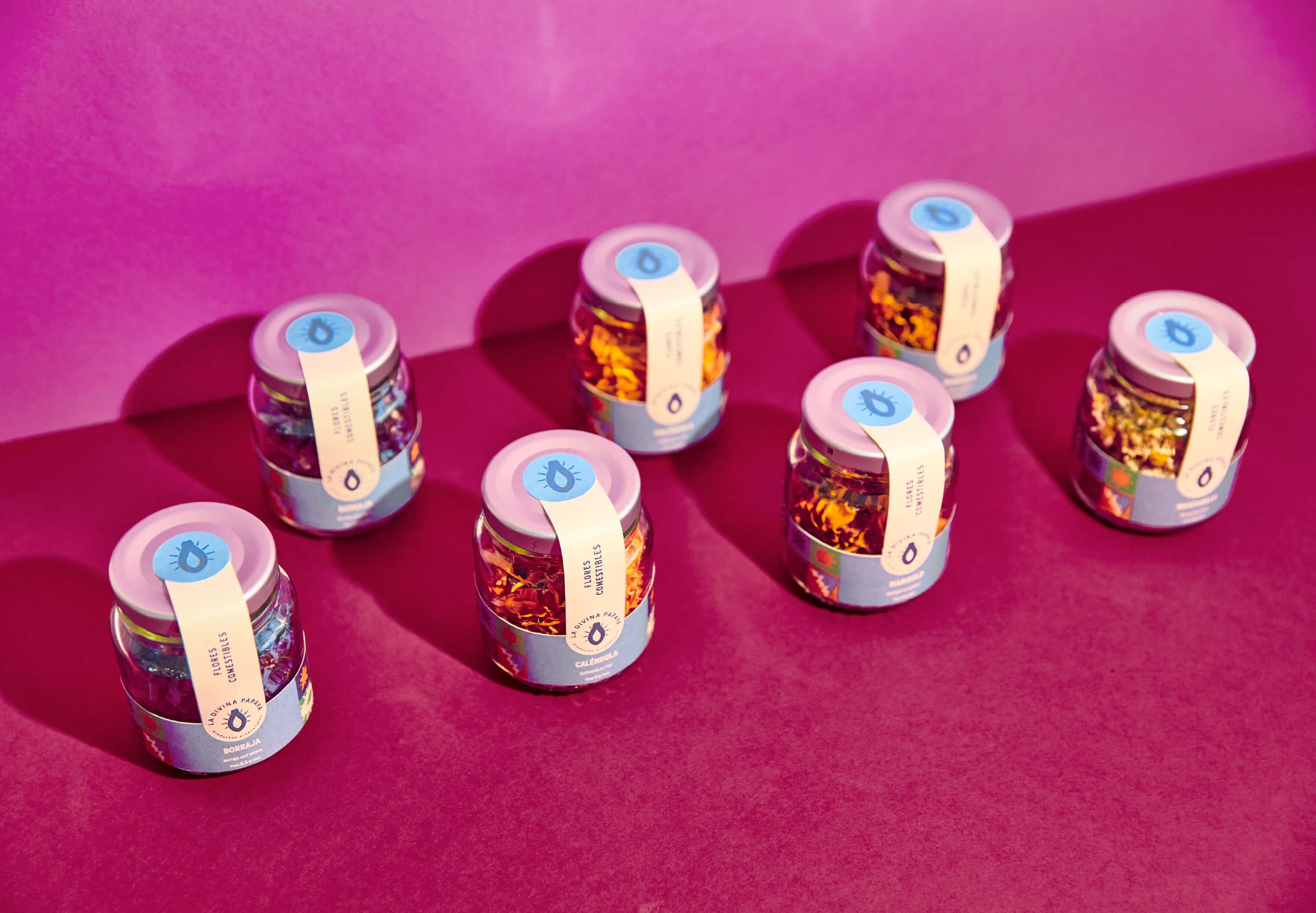 Edible flower jars for La Divina Papaya by Lunalunares, displayed on a bright magenta background with branded labels, circular seals, and colorful artisanal packaging details.
