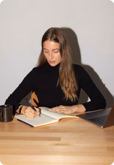 Woman in black sweater writing in a notebook at a wooden desk with a laptop and coffee mug.
