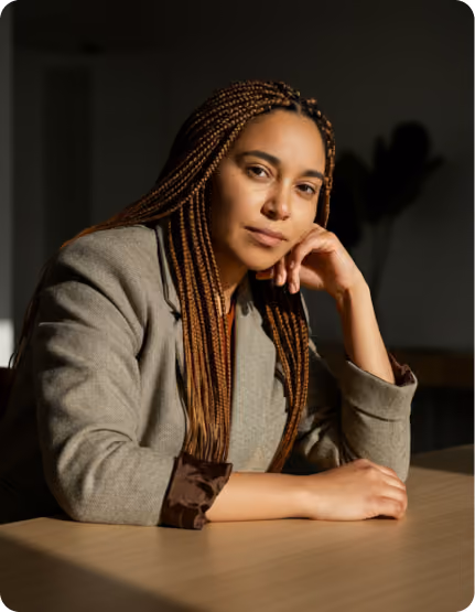 Woman with long braided hair wearing a gray blazer sitting at a table resting her head on her hand.