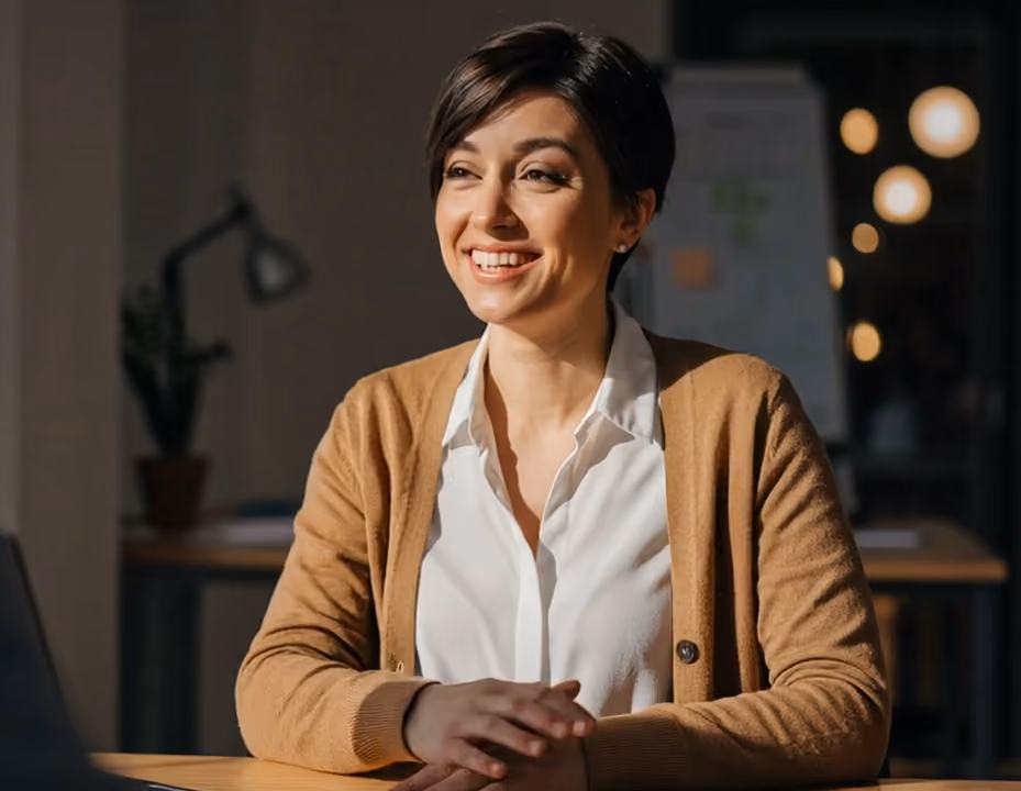 Smiling woman with short dark hair wearing a white shirt and brown cardigan sitting at a desk.