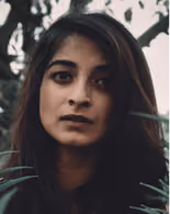 Portrait of a woman with long dark hair and intense expression among green leaves outdoors.