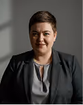 Portrait of a person with short dark hair wearing a black blazer and gray shirt against a plain background.