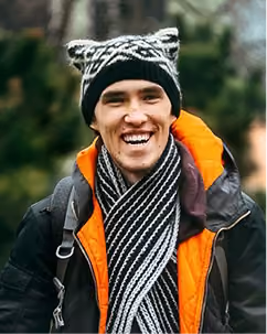Smiling man outdoors wearing a cat-ear knit hat, black jacket with orange lining, and striped scarf.