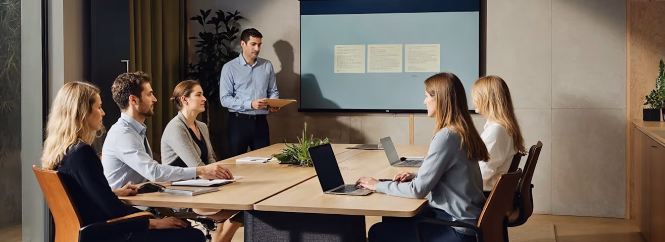 Six professionals in a meeting room with laptops and notes listening to a standing presenter by a screen.