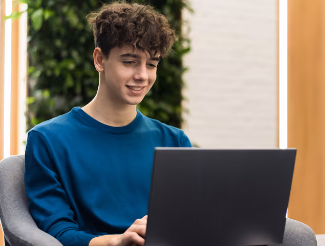 Young man with curly hair in a blue shirt smiling while using a laptop indoors.
