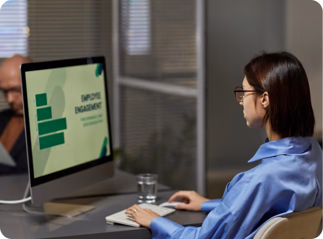 Woman in glasses watching an employee engagement presentation on a desktop computer at an office desk.
