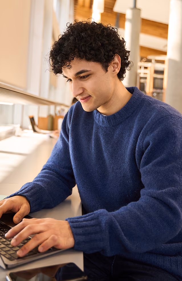 Young man with curly hair in a blue sweater typing on a laptop in a bright room.