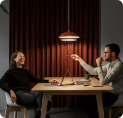 Two people sitting at a table facing each other, talking and laughing in a dimly lit room.