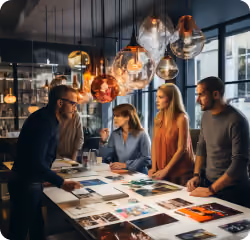 Four people collaborate around a table covered with papers and photos under hanging pendant lights.