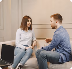 Man and woman sitting on a couch having a serious conversation in a softly lit room.