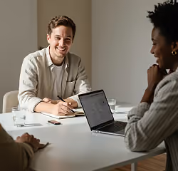 Man smiling and holding a notebook while talking with two colleagues at a table with a laptop.