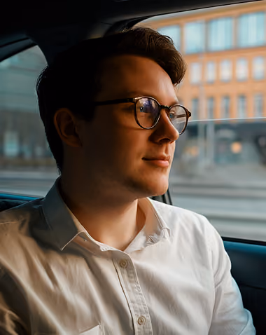 Young man wearing glasses and white shirt sitting in a car looking out the window.